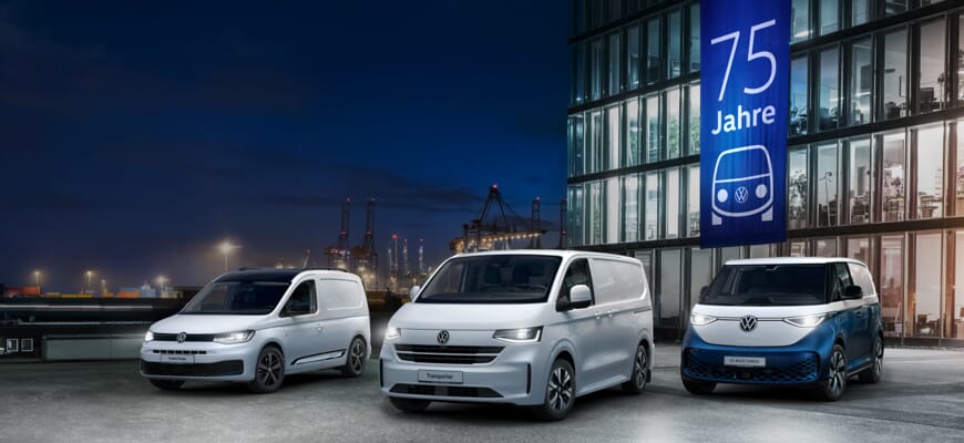 Volkswagen Caddy Cargo, Transporter und ID. Buzz Cargo bei Nacht vor einem beleuchteten Gebäude mit Banner zum 75-jährigen Bulli-Jubiläum.