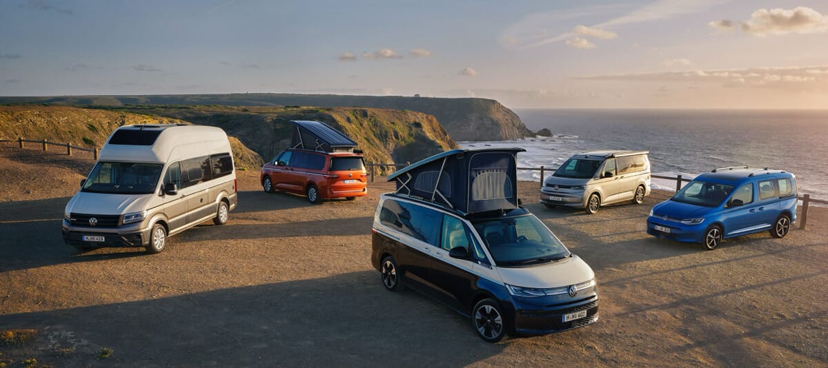 Verschiedene Volkswagen Freizeit- und Reisefahrzeuge auf einer Klippe mit Meerblick bei Sonnenuntergang: Von links nach rechts, Grand California, California in Orange, California in Blau, California in Silber und ein blauer Caddy. Die Fahrzeuge sind teilweise mit ausgeklapptem Aufstelldach zu sehen.