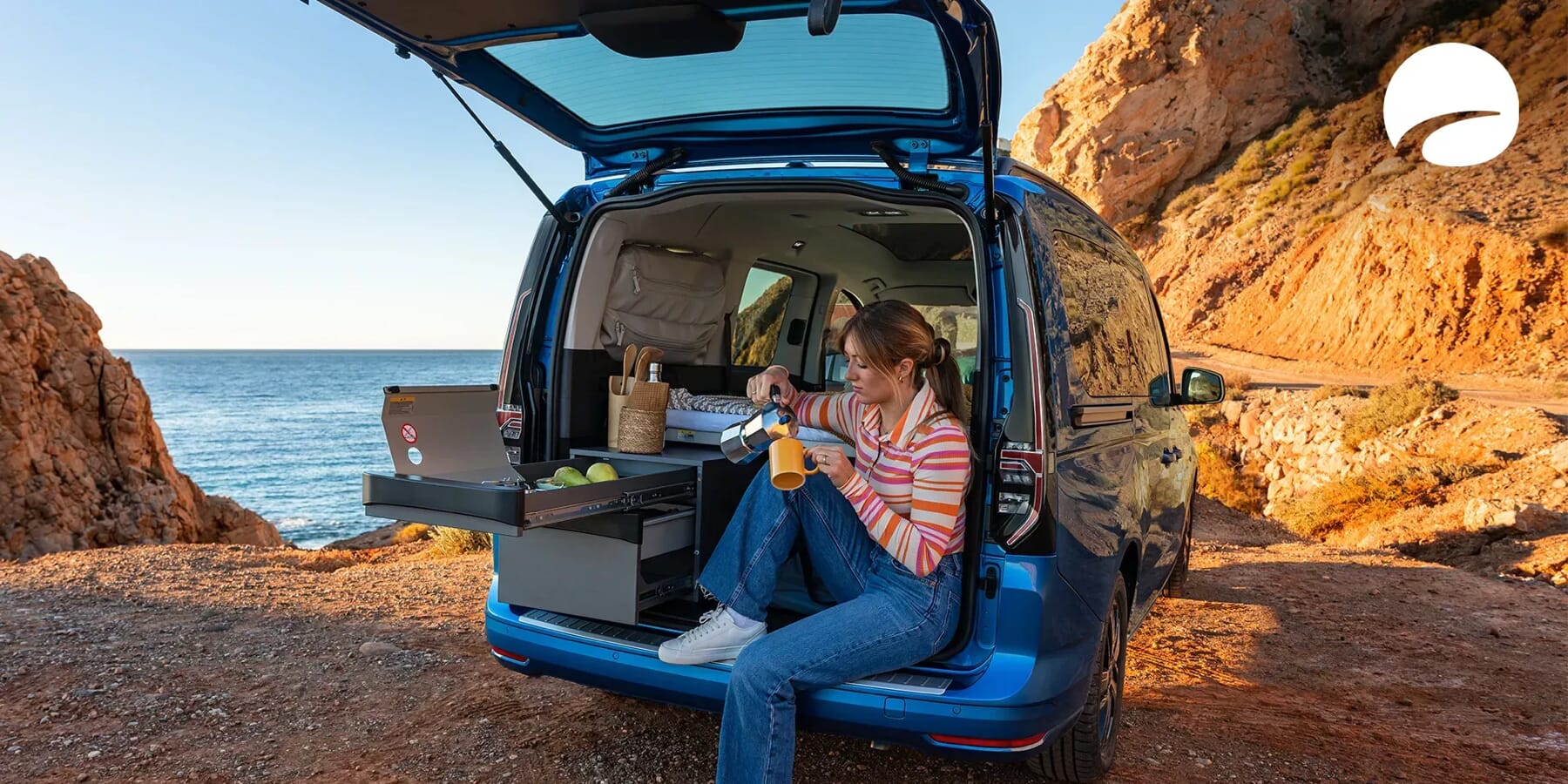 Frau sitzt am Heck eines blauen Volkswagen Caddy California am Meer und nutzt die ausgezogene Campingküche beim Sonnenuntergang in felsiger Küstenlandschaft.