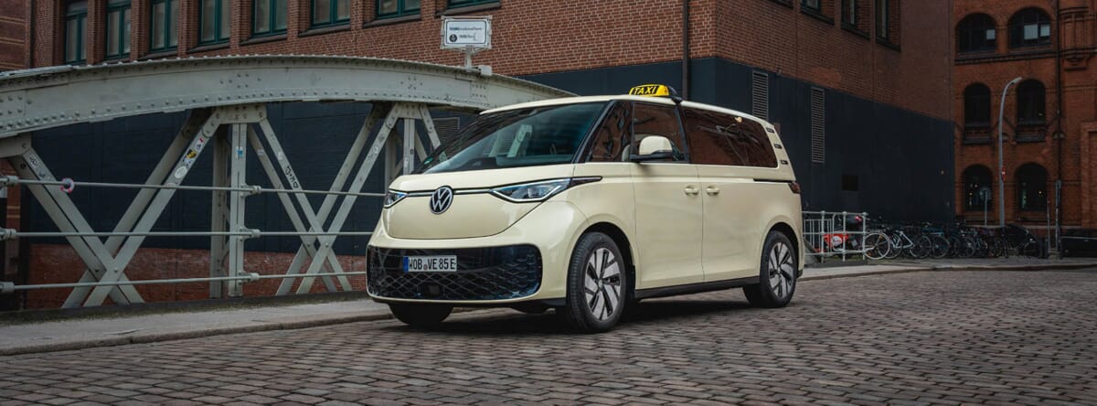 Seitlicher Aufnahme der Volkswagen ID. Buzz E-Taxi mit Industriegebäuden im Hintergrund Das Fahrzeug stehe auf einer Eisenbrücke.