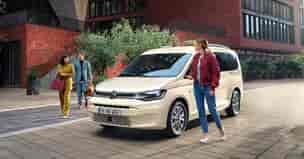 Volkswagen Caddy als Taxiausführung vor einem Backsteingebäude, wo eine Taxifahrerin einen weiblichen und einen männlichen Gast begrüßt. 