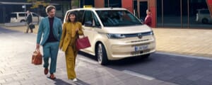 Zwei Passagiere laufen von einem VW Multivan-Taxi nach dem Aussteigen in Richtung Kamera.