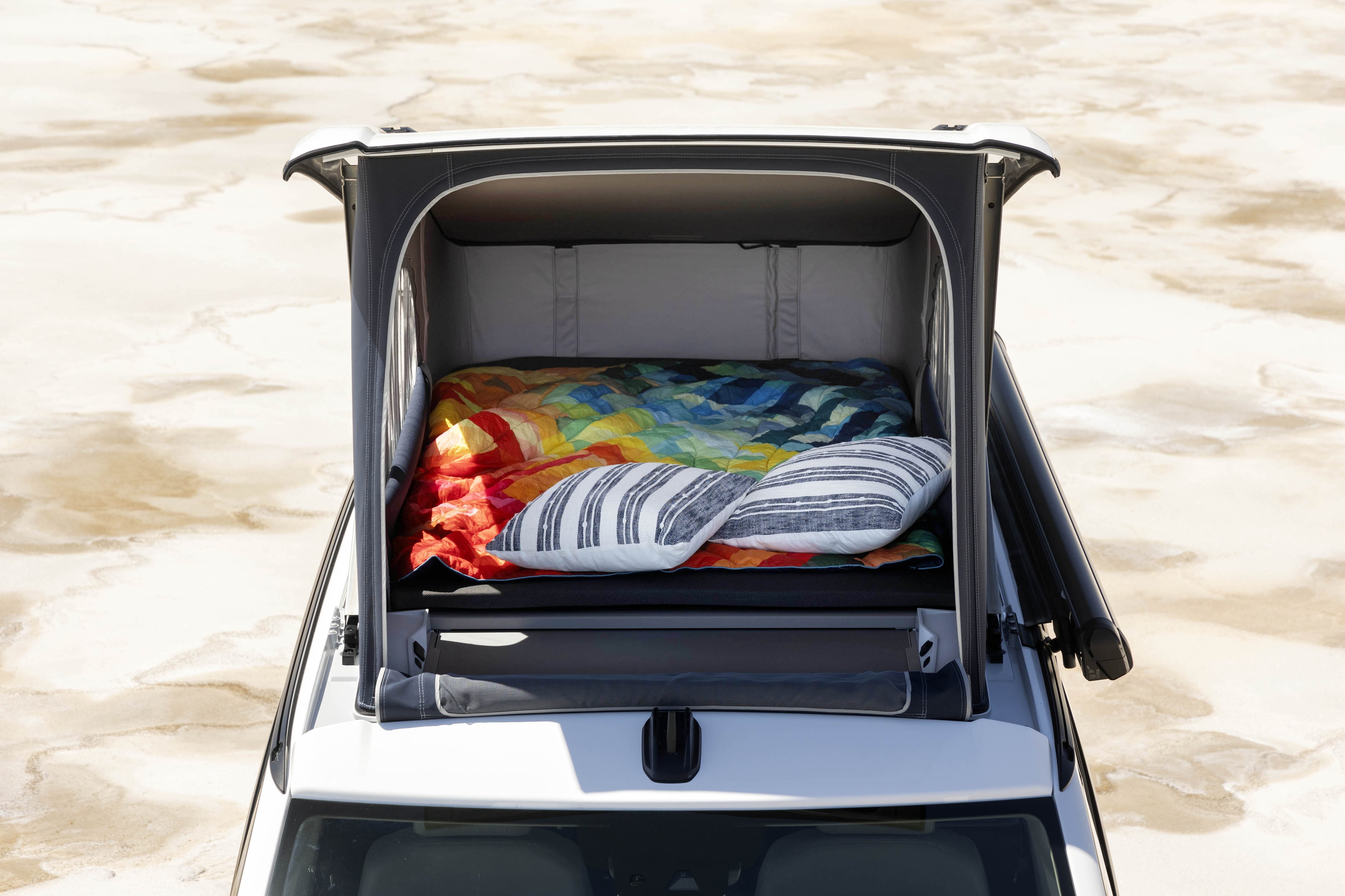 Ansicht von oben des neuen Volkswagen California mit Aufstelldach, buntem Quilt und gestreiften Kissen.