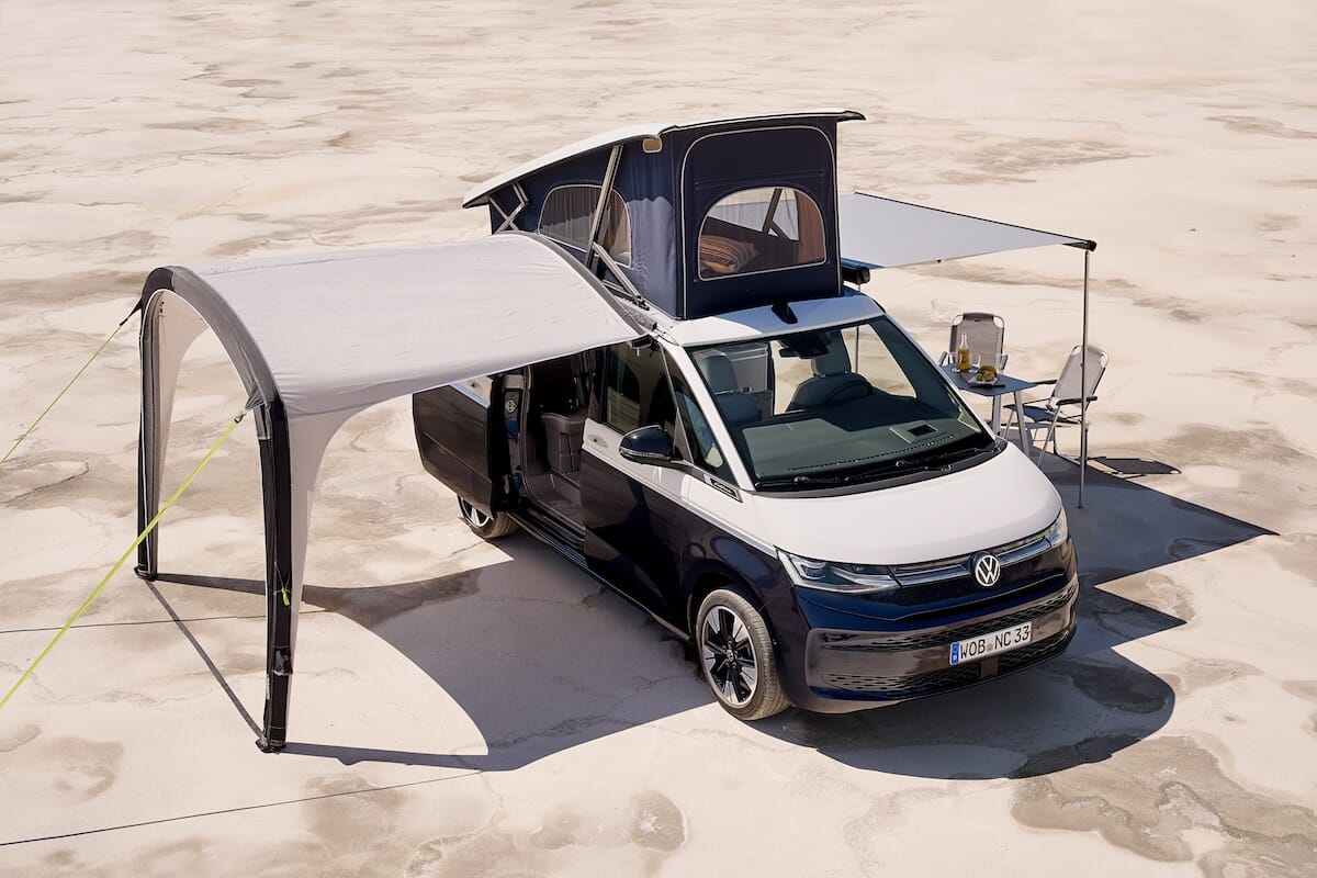 Neuer Volkswagen California mit Aufstelldach und ausgefahrener Markise, eingerichtet mit Tisch und Stühlen auf sandigem Untergrund.