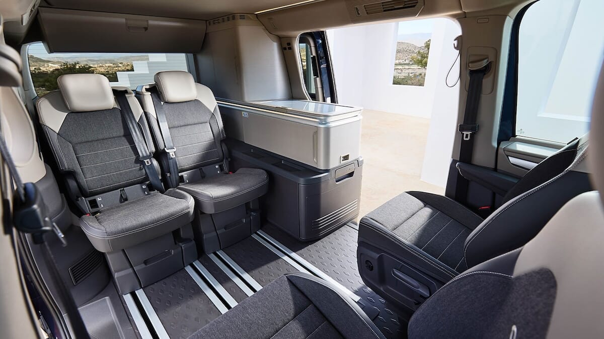 Innenraum des neuen Volkswagen California mit grauen Sitzen, einem eingebauten Stauraum und einer offenen Schiebetür nach draußen.