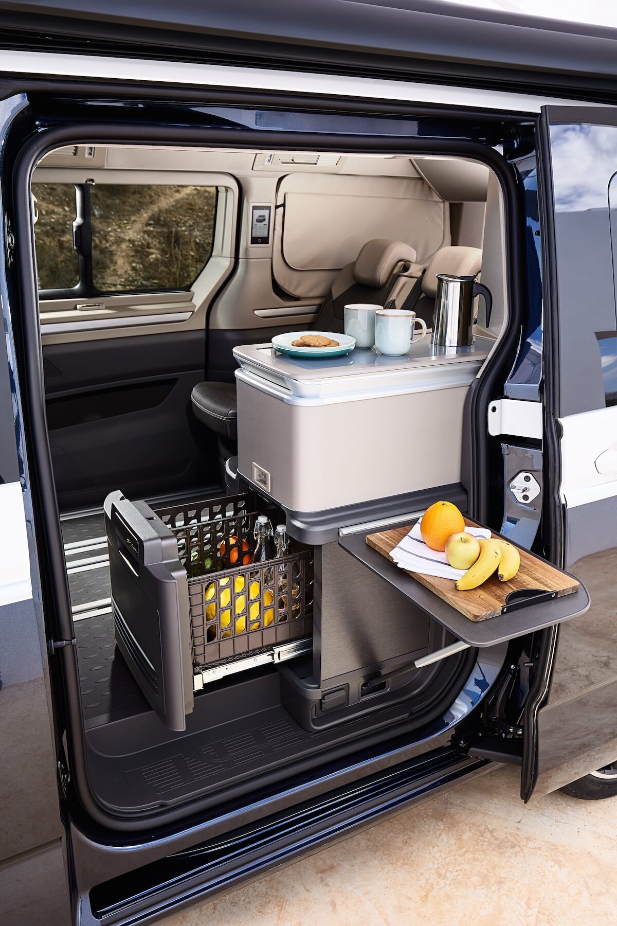 Küchenbereich des neuen Volkswagen California mit offener Schiebetür, zeigt einen Kühlschrank, ein Schneidebrett mit Obst und einen Tisch mit Tassen und Keksen.