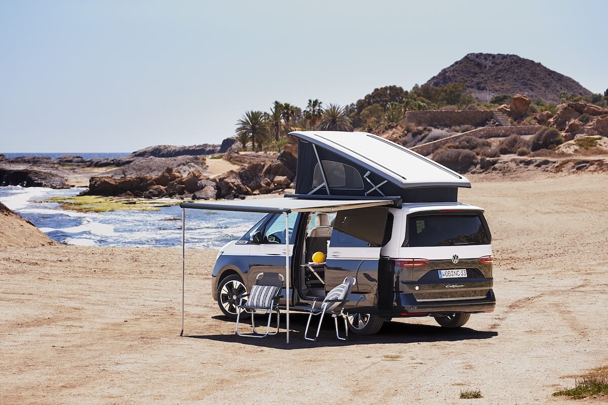 Neuer Volkswagen California mit Aufstelldach und ausgefahrener Markise, eingerichtet mit Stühlen an einem Strand in der Nähe des Ozeans.