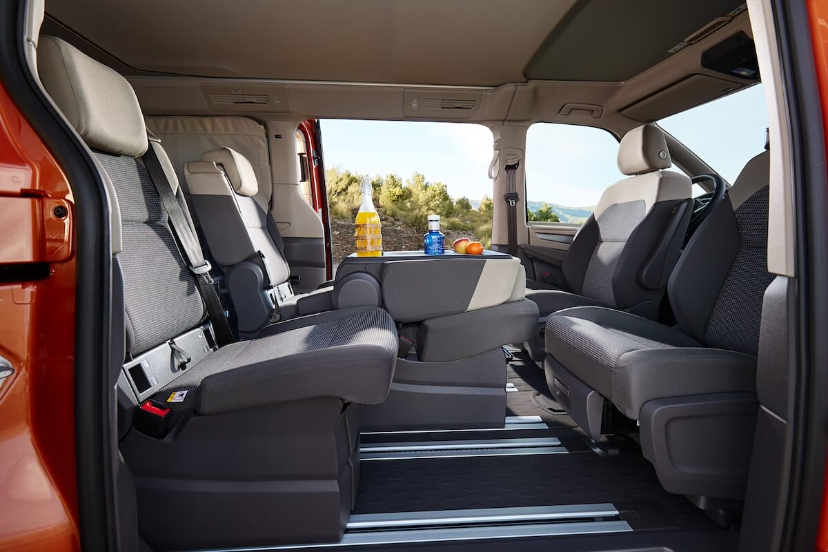 Innenraum des neuen Volkswagen California mit zwei Schiebetüren, grauen Sitzen und einem ausklappbaren Tisch mit einer Flasche Orangensaft, einer Wasserflasche und zwei Orangen.