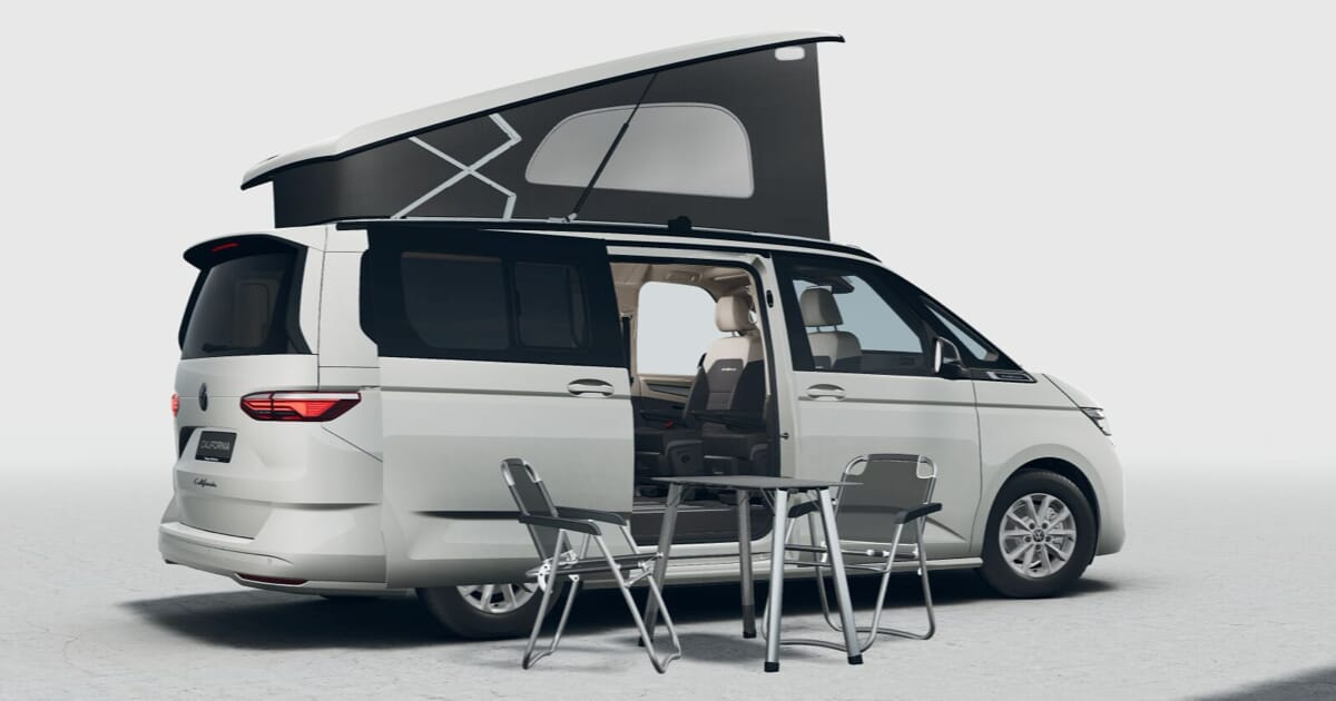 Neuer weißer Volkswagen California T7 2024 mit Aufstelldach, offener Schiebetür und einem Tisch mit Stühlen draußen.