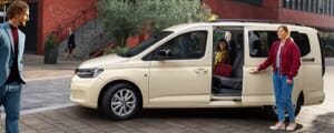 Volkswagen Caddy als Taxiausführung vor einem Backsteingebäude, wo eine Taxifahrerin einen weiblichen und einen männlichen Gast begrüßt.