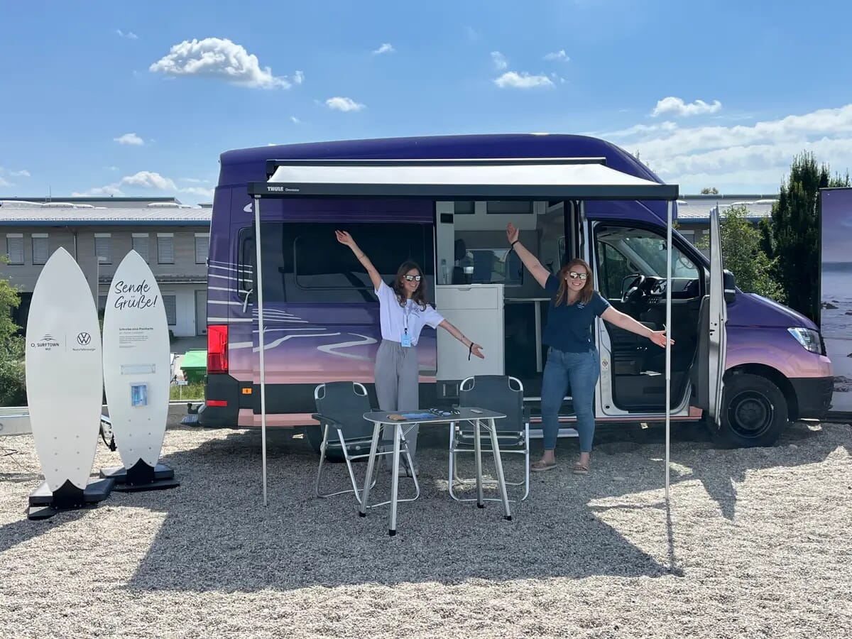 Zwei Frauen posieren mit ausgestreckten Armen vor einem Volkswagen Grand California auf der O2 Surftown MUC Veranstaltung, mit Surfboards in der Nähe ausgestellt und ausgefahrenem Sonnendach des Vans.