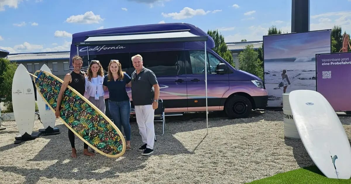 Volkswagen Grand California auf der O2 Surftown MUC Veranstaltung, mit vier Personen, die vorne posieren und ein Surfbrett halten, und weiteren Surfboards in der Nähe ausgestellt.