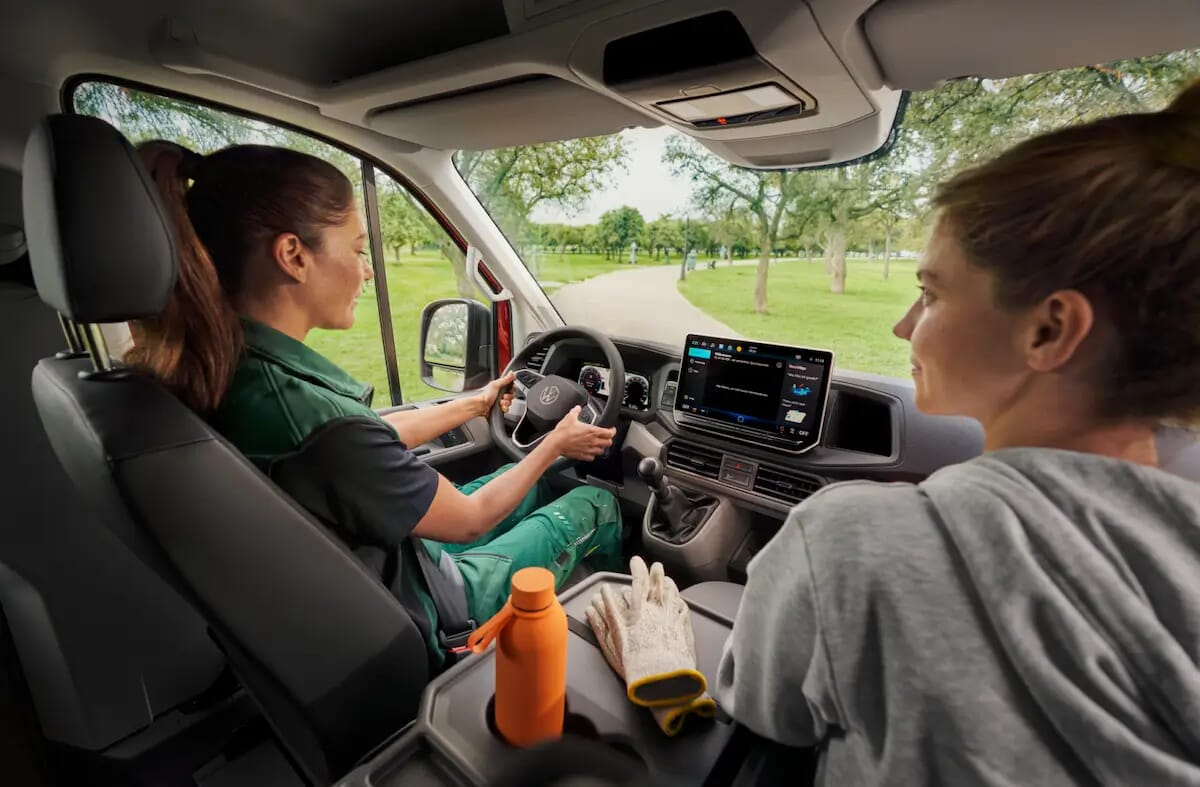 Innenraum eines Volkswagen Crafter mit einer Frau am Steuer und einer weiteren Frau auf dem Beifahrersitz, mit Blick auf das Armaturenbrett und das Infotainmentsystem.