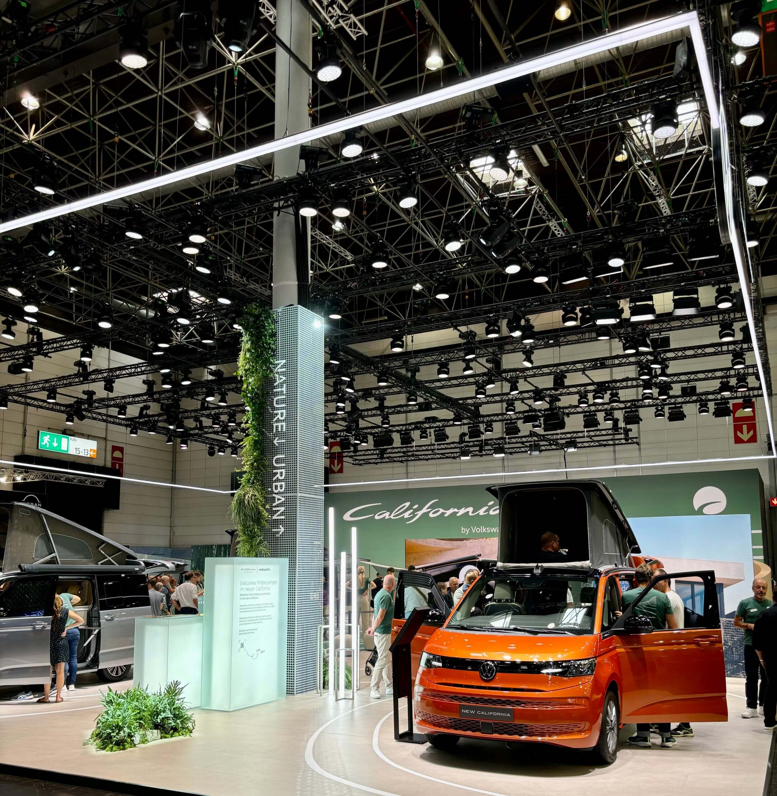 Volkswagen-Stand auf dem Caravan Salon Düsseldorf 2024, mit einem orangefarbenen Volkswagen California mit geöffnetem Aufstelldach, umgeben von Besuchern und einem ‘Nature Urban’-Thema