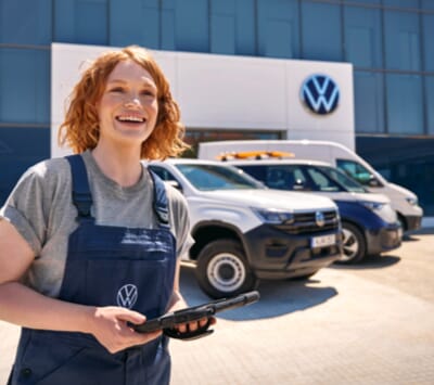 Lächelnde weibliche Volkswagen-Mechatronikerinnen vor einem Volkswagen-Autohaus, die ein Tablet hält, mit zwei geparkten Volkswagen-Nutzfahrzeugen im Hintergrund.