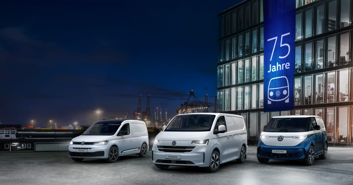Drei weiße Volkswagen Nutzfahrzeuge – Caddy Cargo, Transporter und ID. Buzz Cargo – stehen bei Nacht vor einem modernen Gebäude mit einem Banner zum 75-jährigen Jubiläum des VW Bulli.