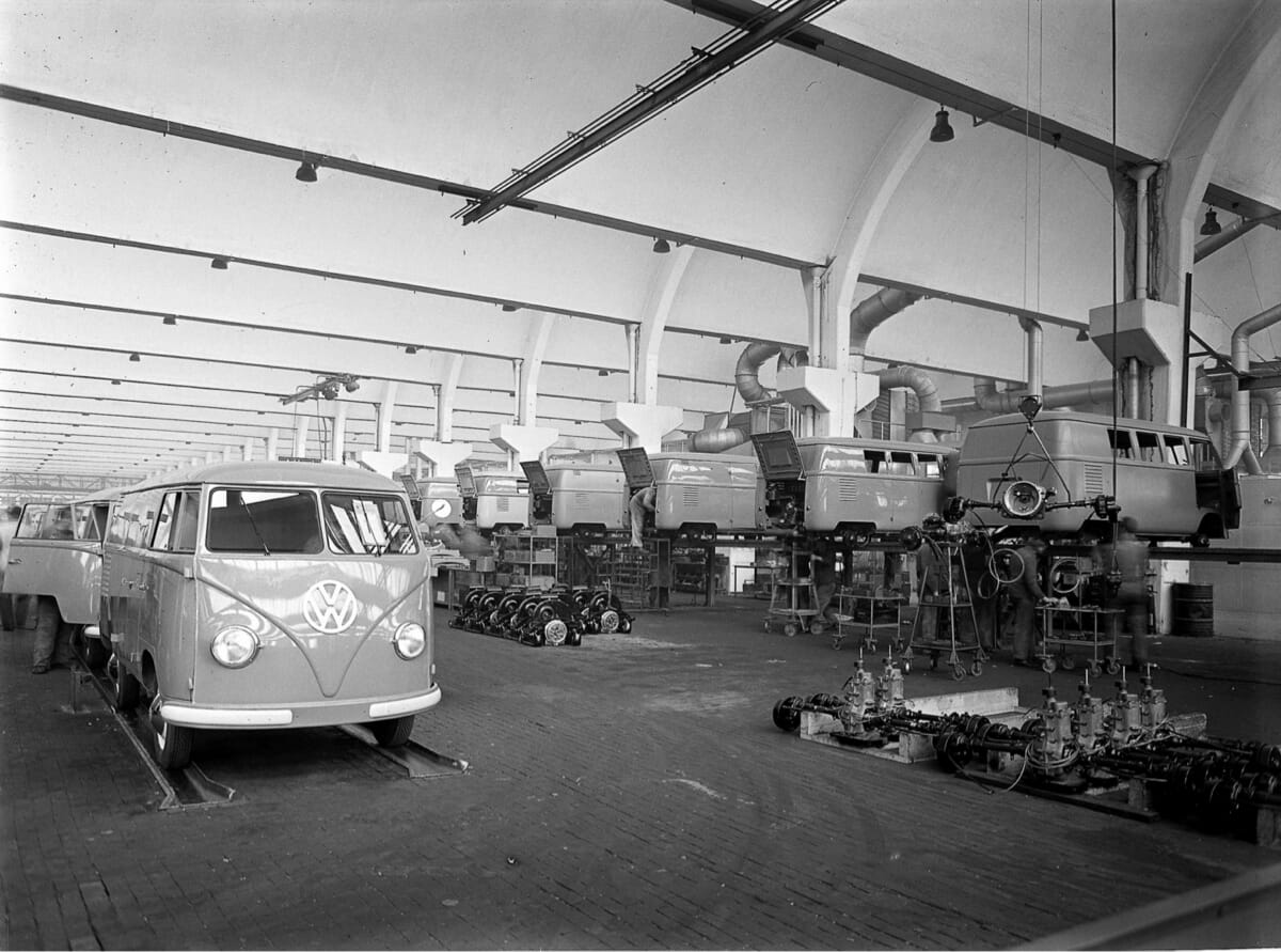 Historische Schwarz-Weiß-Aufnahme der Volkswagen Transporter Produktion in Wolfsburg im Jahr 1950, mit mehreren VW Bullis in der Montagehalle.