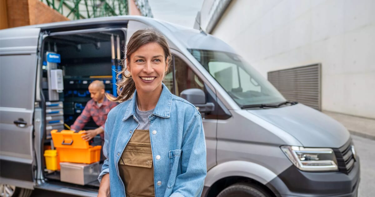 Lächelnde Handwerkerin vor einem silbernen Volkswagen Crafter mit geöffneter Seitentür, im Hintergrund ein Kollege.