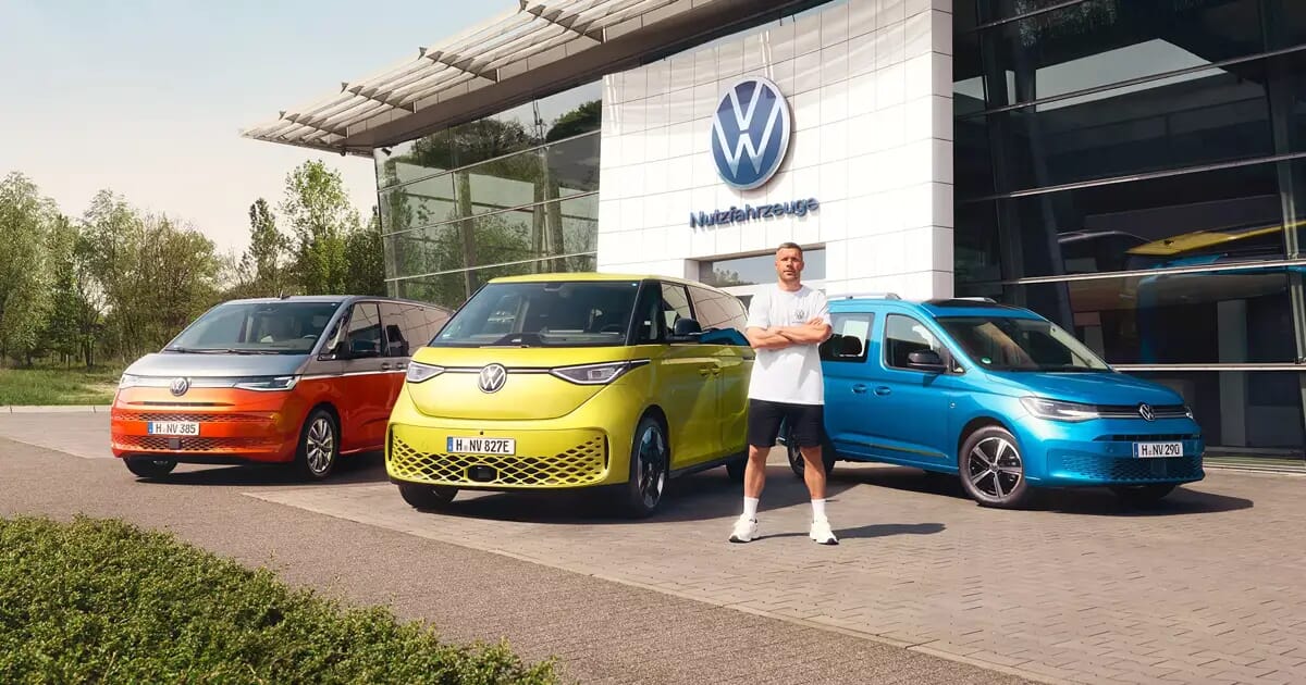 Volkswagen Energy Sondermodelle vor einem VW Autohaus: orangefarbener Multivan, gelber ID. Buzz und blauer Caddy, mit Lukas Podolski stehend im Vordergrund.