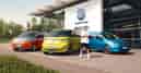 Volkswagen Energy Sondermodelle vor einem VW Autohaus: orangefarbener Multivan, gelber ID. Buzz und blauer Caddy, mit Lukas Podolski stehend im Vordergrund.