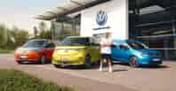 Volkswagen Energy Sondermodelle vor einem VW Autohaus: orangefarbener Multivan, gelber ID. Buzz und blauer Caddy, mit Lukas Podolski stehend im Vordergrund.