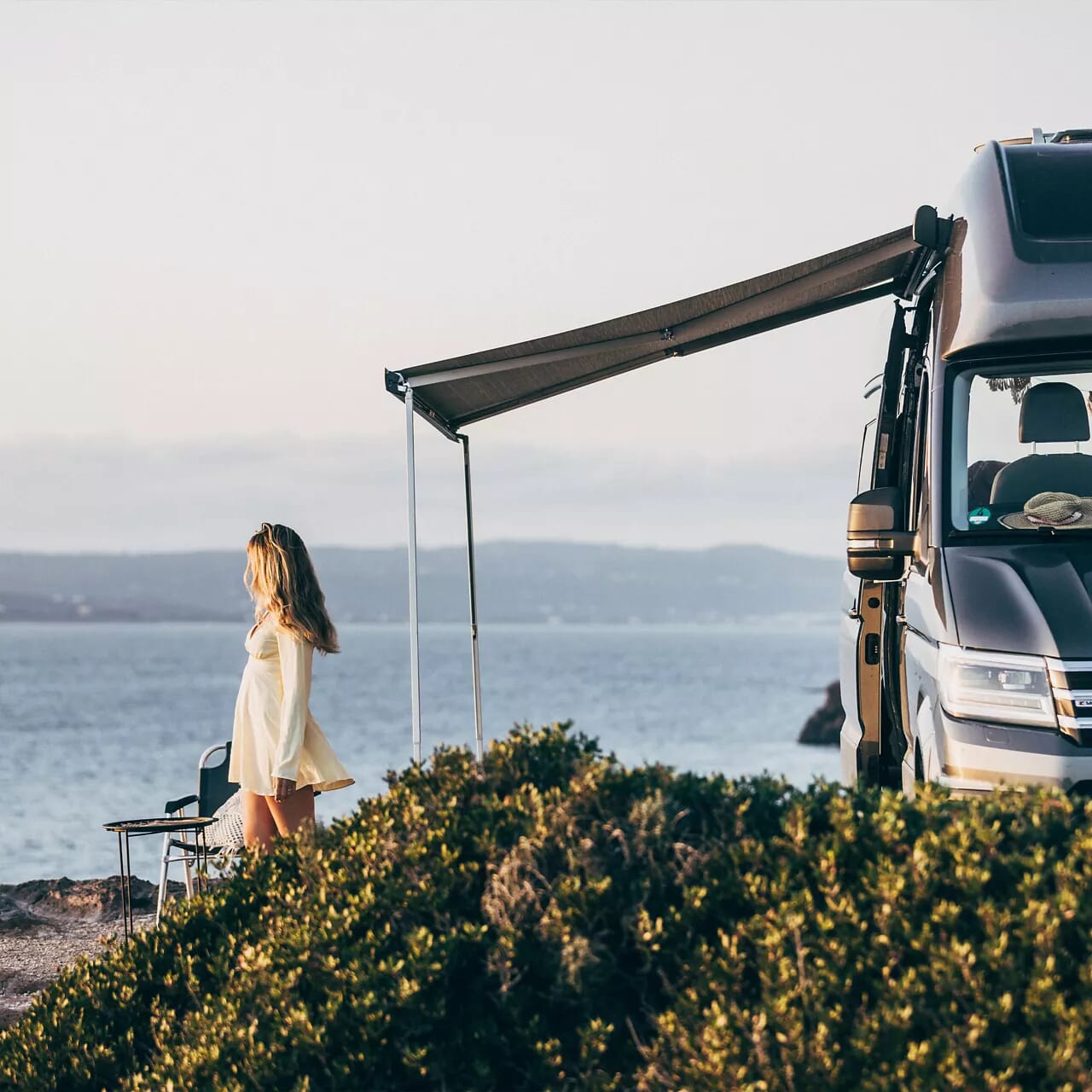 VW Grand California mit ausgefahrener Markise und Campingmöbeln an der Küste.