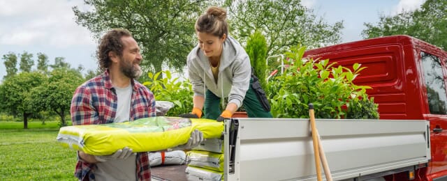Mann und Frau laden Gartenzubehör, darunter Pflanzen und Säcke mit Erde, auf die Ladefläche eines roten Volkswagen Transporter Pritschenwagen in einer grünen Außenumgebung.