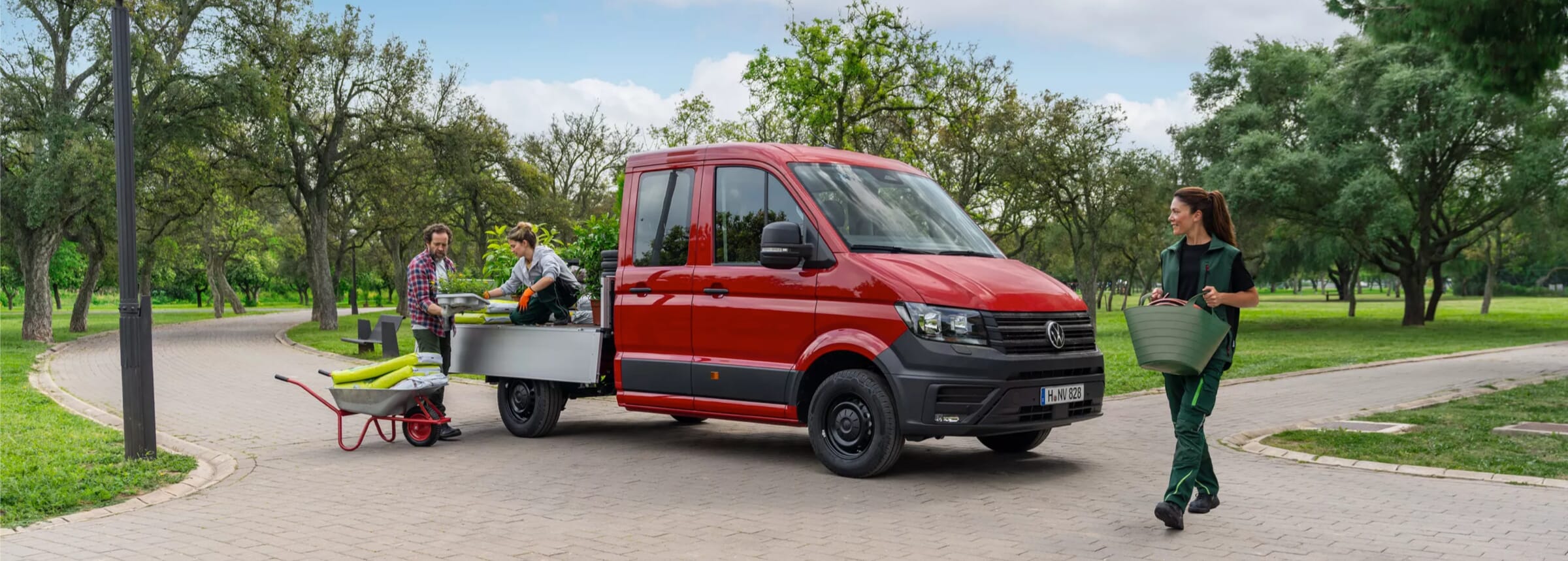 Personen laden Gartenzubehör auf die Ladefläche eines roten Volkswagen Crafter in einem Park, während eine Person in der Nähe einen grünen Eimer trägt.